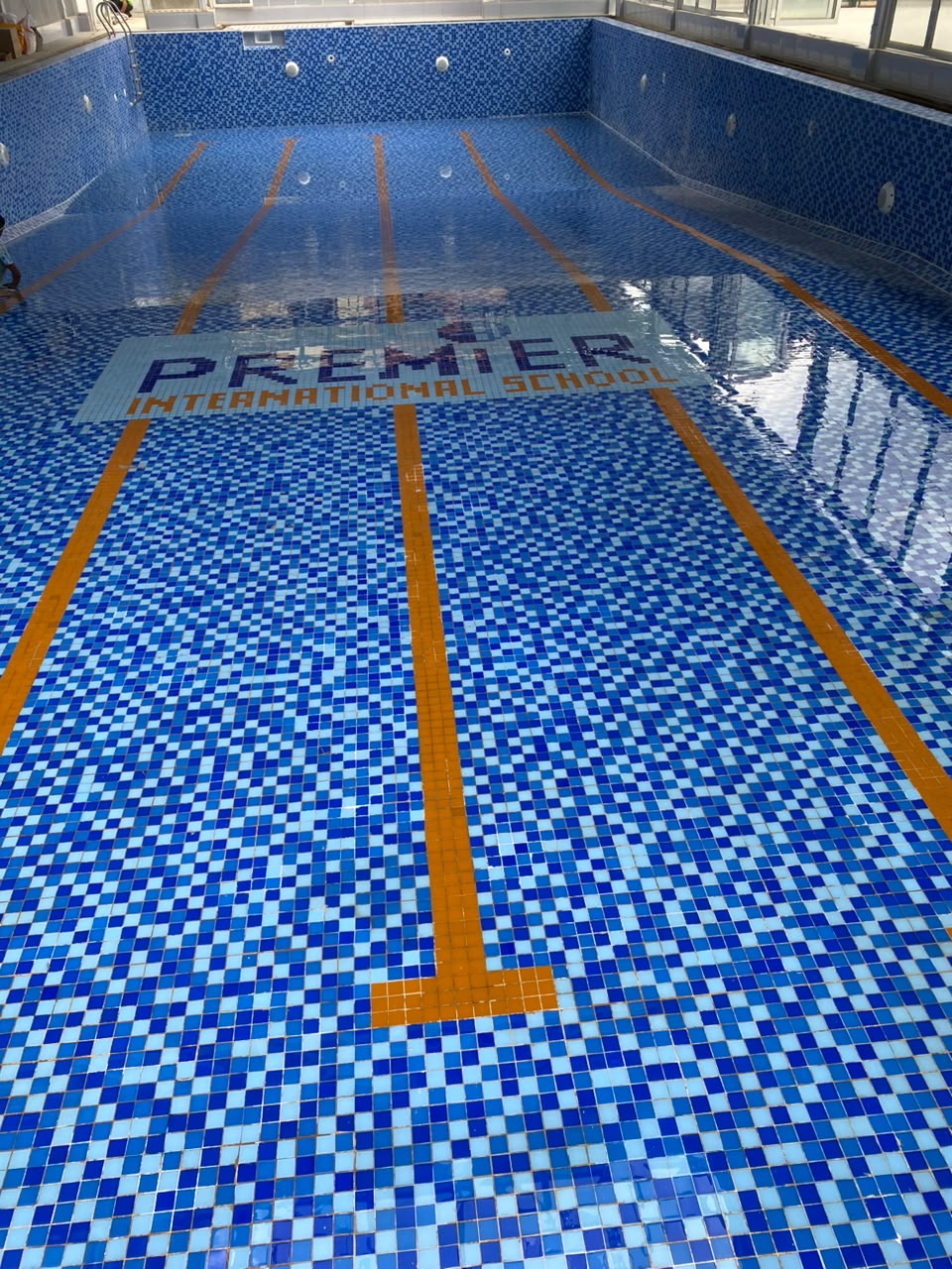 Blue mosaic pool floor at Premier International School, with the school name and yellow swimming lane markings.