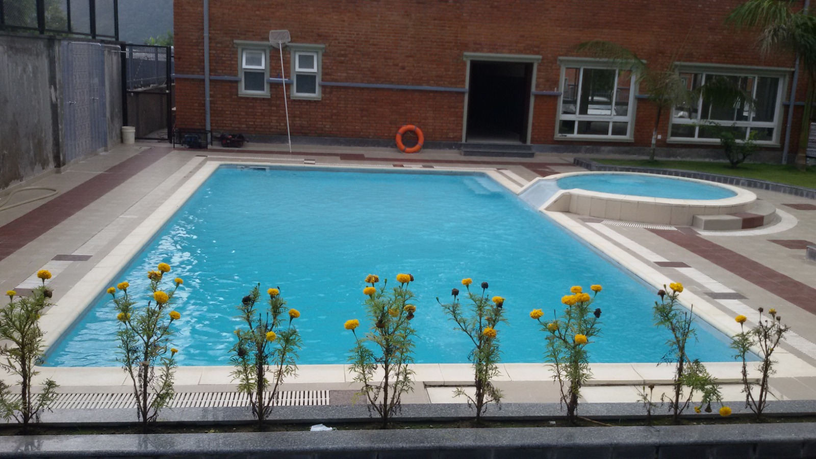 Rectangular swimming pool and adjoining round smaller pool at Deep Housing, with marigolds and a brick building beside the deck.