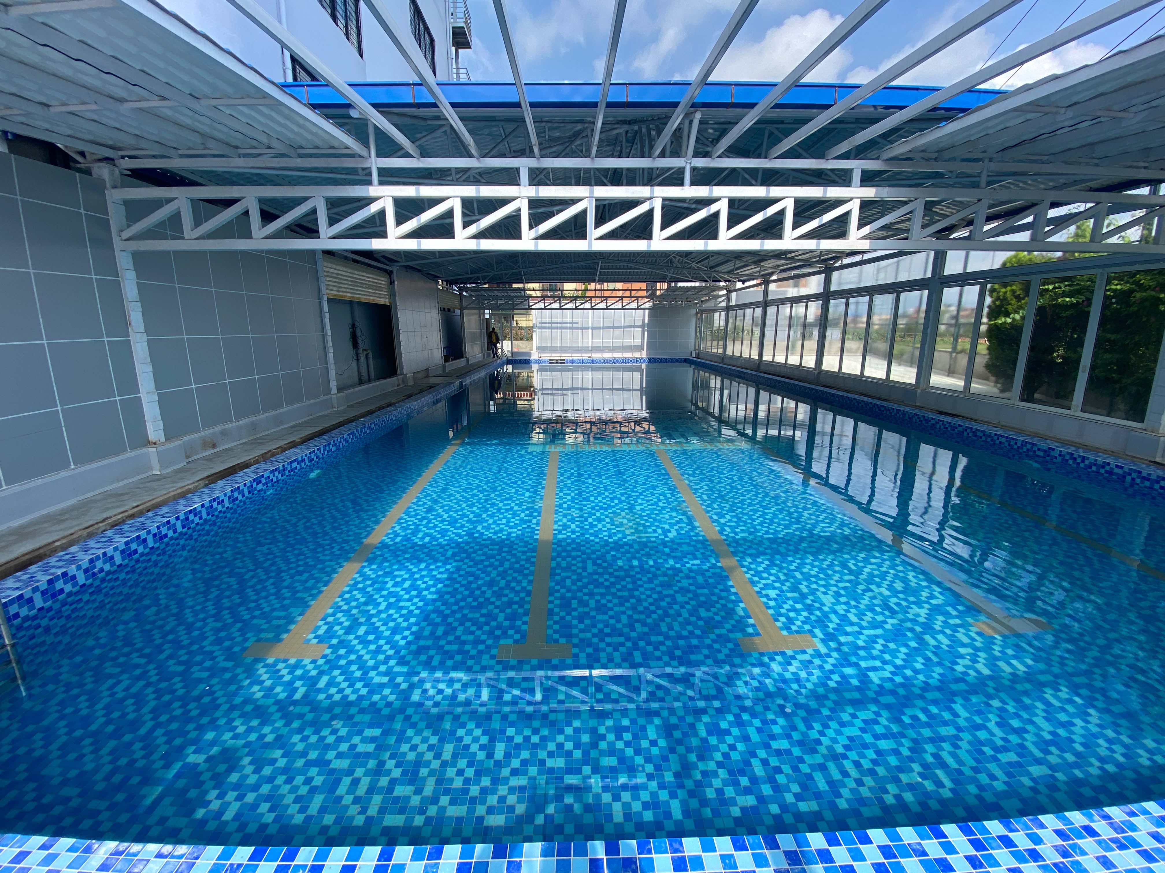 Covered swimming pool at Premier International School, with blue mosaic tiles, yellow lane markings and a steel-framed roof.