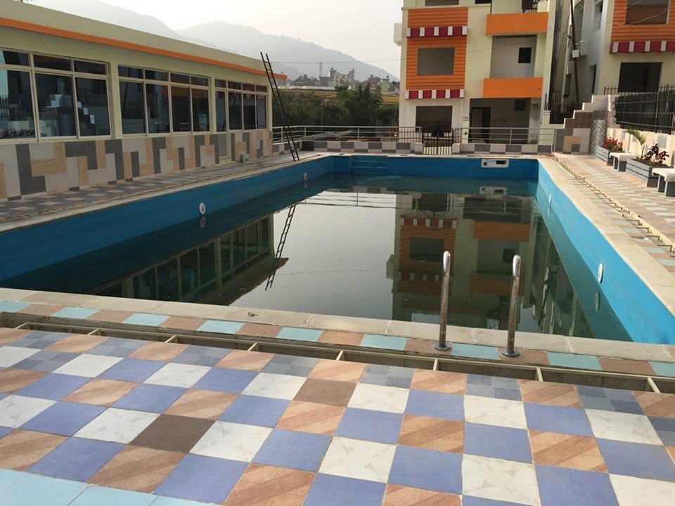 Rectangular swimming pool at Padma Colony, with entry steps, a stainless-steel ladder, a tiled deck and surrounding residential buildings.
